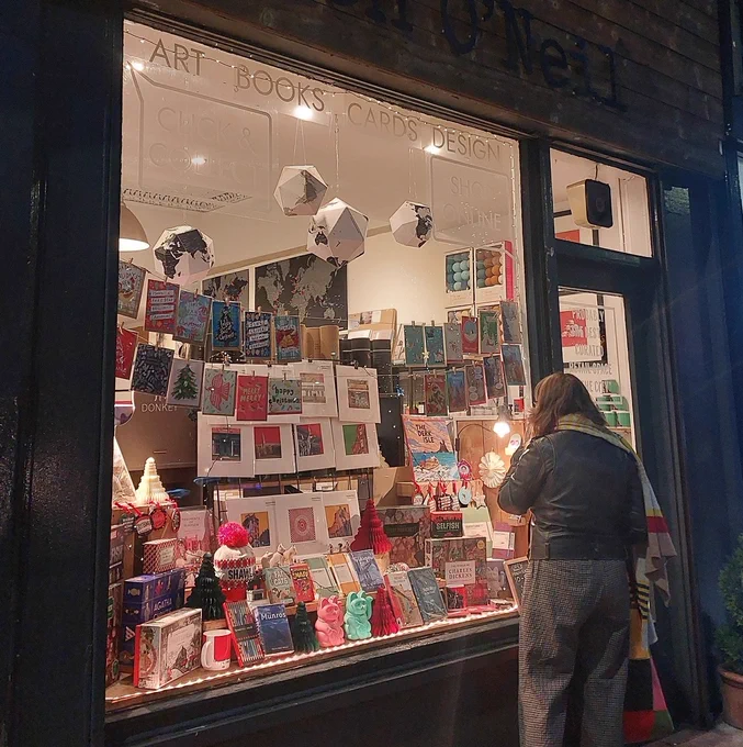 Shop window display at Stephen O'Neil's store in Shawlands featuring a range of art prints, cards, books, games, models and toys, knitwear, homeware, and more.
