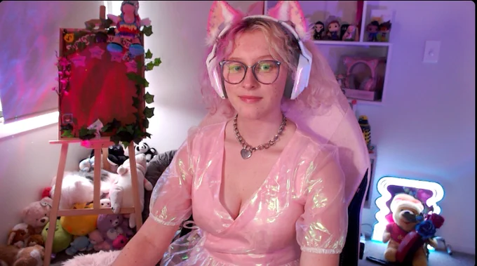 a photo of a girl with pink and white hair and a white headset with pink cat ears, sitting in a pink chair with a shiny pink dress, and a chunky silver necklace. 