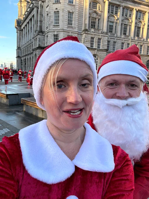 The image shows a close-up selfie of two people dressed in Santa Claus outfits. The woman in the foreground is wearing a red Santa hat with a white trim and a red velvet outfit with a white collar. She has blonde hair and is smiling. Behind her, a man is also dressed in a Santa outfit, with a red hat and a white beard. In the background, there are several other people dressed in similar Santa outfits, gathered in front of a large, ornate building with many windows. The sky is clear and blue, suggesting a sunny day.