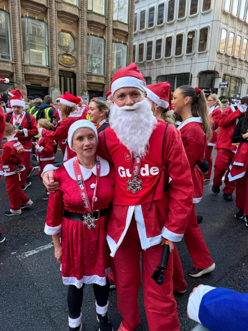 The image shows a festive scene with a group of people dressed in Santa Claus outfits, likely participating in a Santa-themed event or run. In the foreground, there is a man and a woman posing together. The man is wearing a red Santa suit with a white beard and a red hat. His shirt has the word "Guide" printed on it in white letters, and he is holding a small camera on a stick. The woman is wearing a red dress with white trim, a black belt, and a Santa hat. Both are wearing medals with snowflake designs around their necks. In the background, more people are dressed in similar Santa outfits, and the setting appears to be a city street with tall buildings.