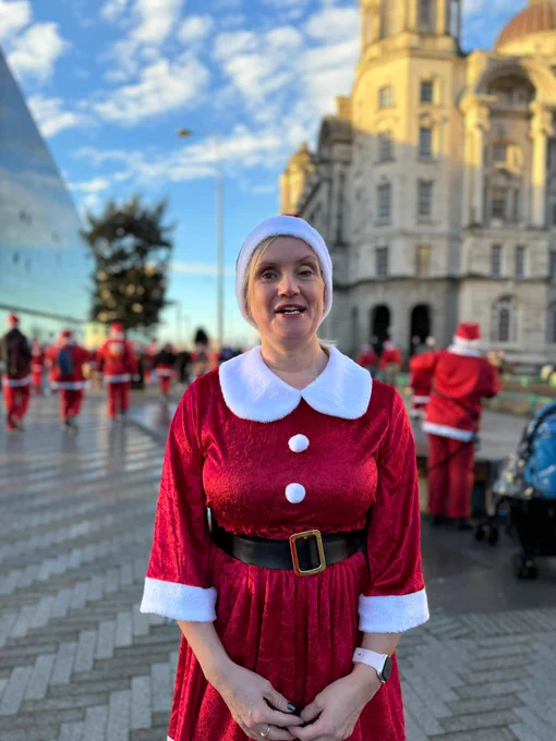 The image shows a woman dressed in a festive Santa Claus outfit, which includes a red velvet dress with white fur trim on the collar and cuffs, and a black belt with a gold buckle. She is also wearing a Santa hat. The background features a group of people, many of whom are also dressed in Santa costumes, suggesting a festive event or gathering. The setting appears to be outdoors in an urban area, with a large historic building on the right and a modern glass building on the left. The sky is blue with scattered clouds.
