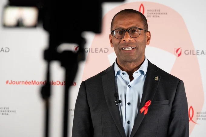 L’honorable Greg Fergus, Président de la Chambre des communes, portant un ruban rouge pour montrer sa solidarité dans la lutte contre le VIH/sida. Derrière lui, un arrière-plan sur lequel figurent les mots « Gilead », « Journée Mondiale du SIDA » et « Société canadienne du sida », et à sa gauche, l’ombre d’un appareil-photo.