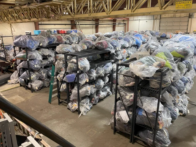 The inside of a large facility. Three rows of large industrial shelves are full of clear garbage bags. A row of shelves in the back has coolers on top of it.