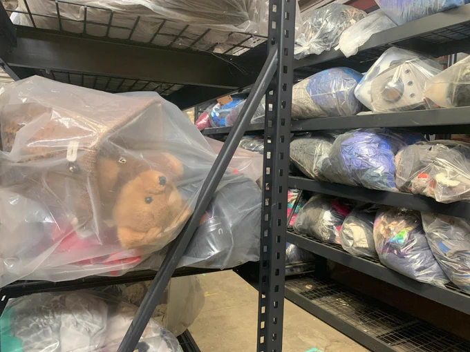 A close-up photo of industrial storage shelves holding clear bags. One bag has a teddy bear inside. Many of the bags in the background have textiles, such as clothing, inside.