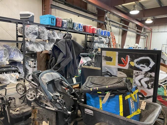 An industrial shelf with coolers on top and clear garbage bags on the shelves below. In front of the shelf are various items, such as a folded up canopy, two strollers, a piece of artwork and a wheelbarrow with bags inside.