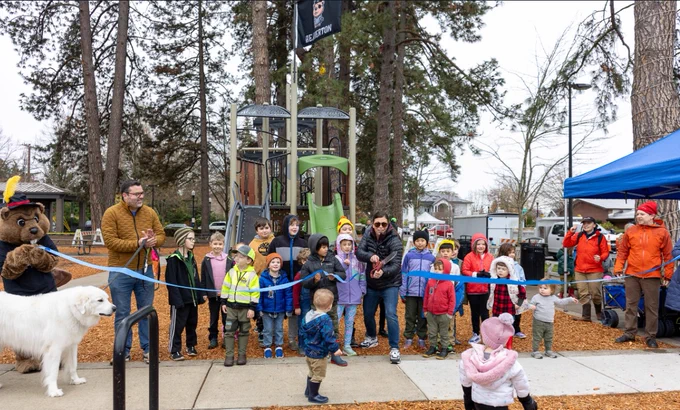 Mayor Lacey Beatty cuts the ribbon in front of a playground structure.  Many children and a few adults are with her as well as Billy the Beaver in a pirate hat.