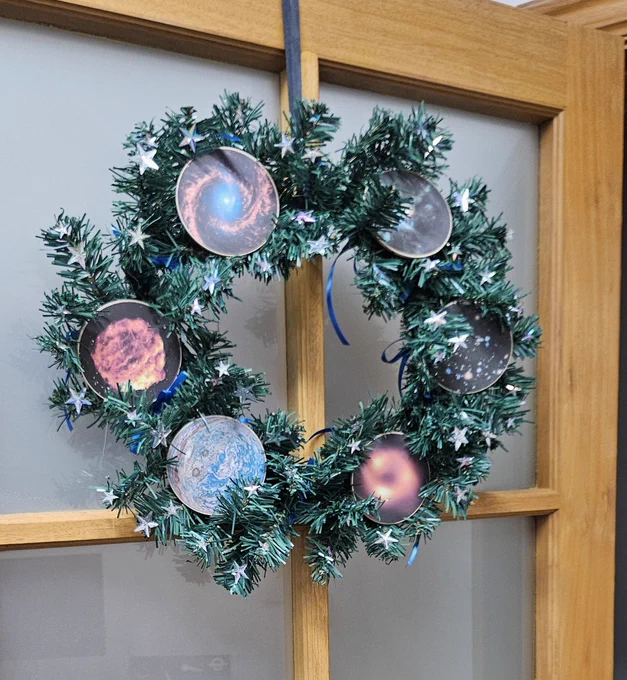 A dark green wreath hangs on a door. The wreath has small, shiny stars on it and 6 circular astronomy images. The images are: a JWST image of a galaxy, a MeerKAT image of the Galactic Centre, the Hubblr Ultra Deep Field, the EHT image of the black hole at the centre of our Galaxy, a Juno image of Jupiter, and an ASKAP image of a supernova remnant