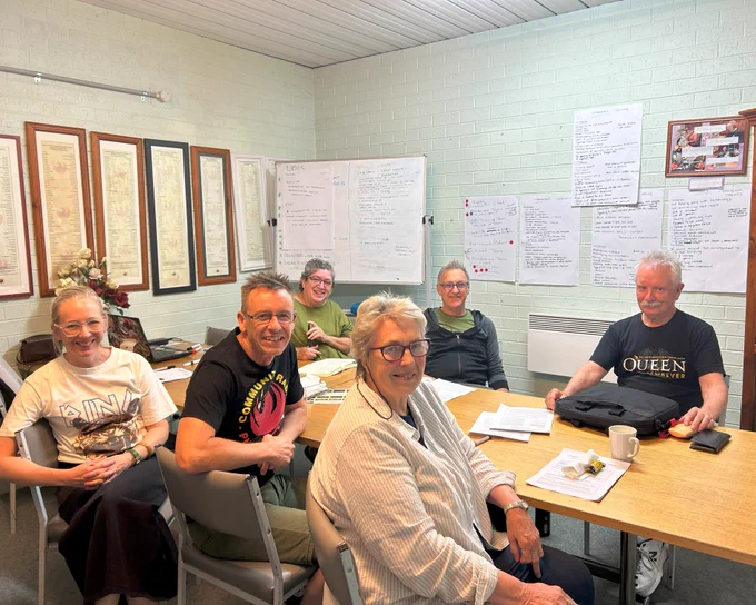 Image of 6 people sitting at a table. On the wall behind them are papers filled with writing and a whiteboard full of writing as well. 