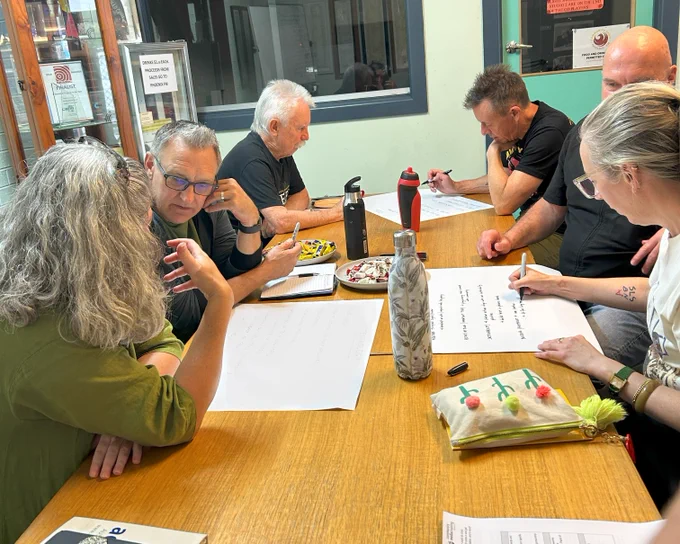 photo of six people sitting at a table. They are conversing and writing on paper.