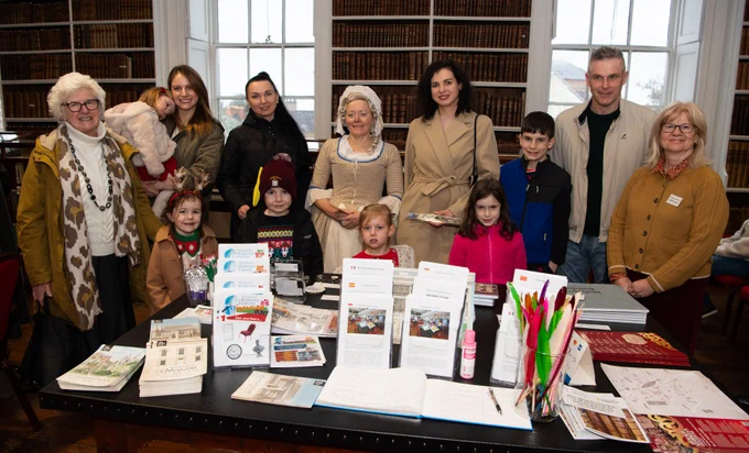 Group of thirteen people of all ages behind a table full of flyers, maps and small shops items in an old library.