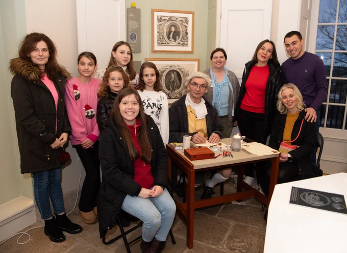 Group of ten adults and children standing and sitting around a living history character in 18th century clothing, who is seated at a table with a wax seal set.