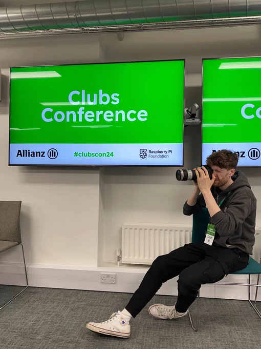Matthew is a young man dressed in a grey Raspberry pi hoodie with black jeans and white trainers. He is sitting on a chair and using a camera with zoom lens to take photos at an event. There is a screen behind him with a green background and white writing that says Clubs Conference in large lettering, and Allianz, #ClubsCon24 and Raspberry Pi Foundation