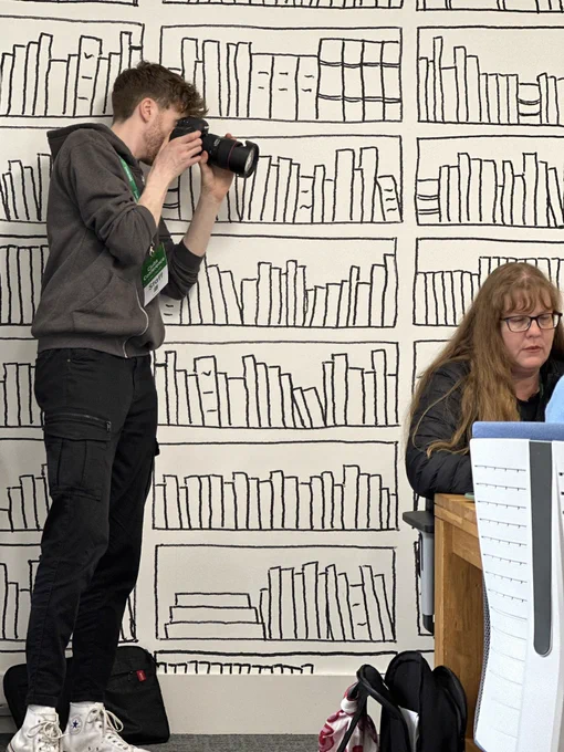 Matthew is a young man dressed in a grey Raspberry pi hoodie with black jeans and white trainers. He is standing in front of a white wall that has line drawings of library shelves. He is using a camera with zoom lens to take photos at an event. To the right of the image is a lady with long hair and glasses . She is using a computer in one of the programming workshops