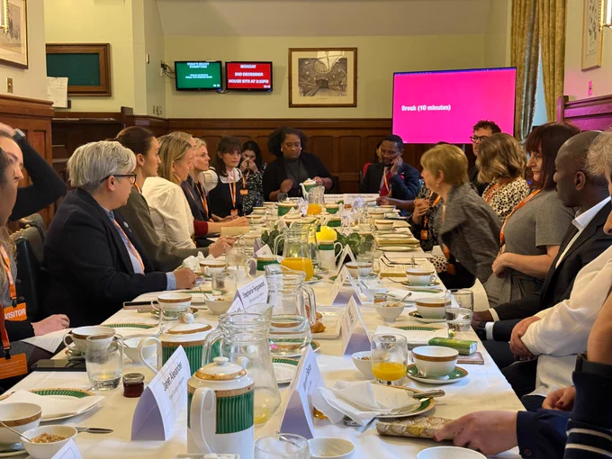 Bell pours tea at the head of a packed table in a parliamentary committee room. There are two screens behind her: one red, one green. Tea, juice and breakfast items are laid out on the table. About a dozen people sit around the table.