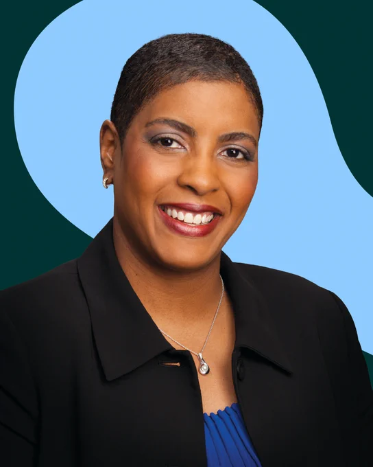 Person with short dark hair smiling, wearing a dark blazer and blue top with a pearl necklace.