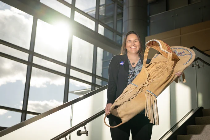 Tami, a woman of Native American heritage from the Delaware Nation, smiles standing on a staircase in a modern, sunlit building, holding a traditional handcrafted cradleboard adorned with intricate beadwork. She is wearing a paisley patterned shirt covered with a black sweater that has the NASA insignia on the right breast. Credit: NASA/Charles Beason