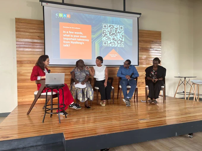 Five speakers sit on stage during a panel discussion. Angeline talks into a microphone while the other panellists listen. 