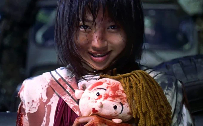 On the right, a still from the film BATTLE ROYALE (2000) in which a preteen Japanese girl in braces, clutching a bloodied doll, smiles in triumph, having won a battle.