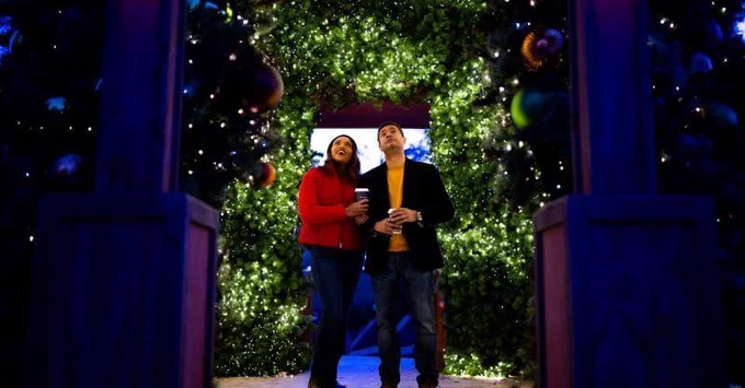 A couple stands in a twinkly room of lights, ornaments, and garland while gazing up with wonder. 