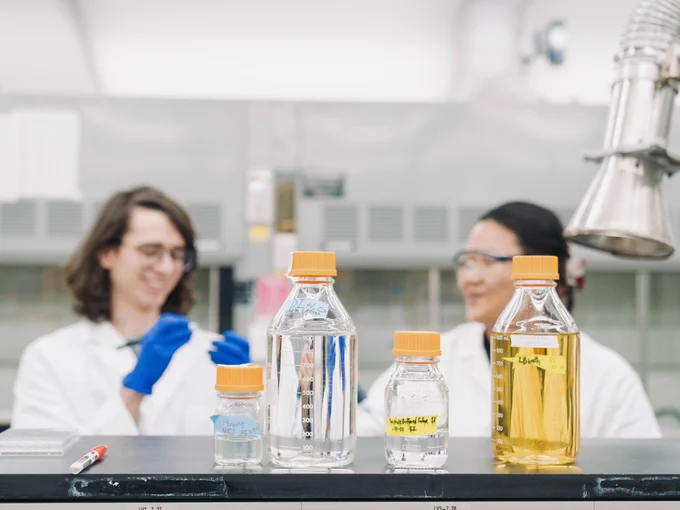 Two laboratory technicians smiling and engaging in a conversation while sitting at a lab bench with several bottles of chemicals in front of them.