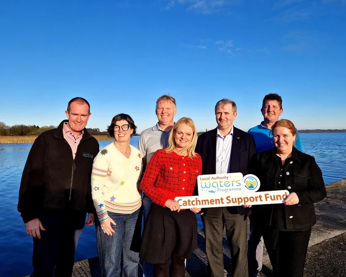 LAWPRO staff standing beside a lake on a sunny day. From left to right: Barry Deane, Margaret Keegan, Dr. Fran Igoe, Sheevaun Thompson, Anthony Coleman, Jimmy McVeigh, Bernadette White. They are holding a sign that reads "Local Authority Waters Programme Catchment Support Fund."