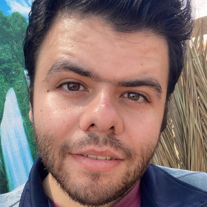 close-up of Khaled outside in the sun, dark brown hair combed up, slight brown beard and hazel-brown eyes staring into the camera. He wears a slight smile and a blue and gray zip up with maroon shirt underneath.