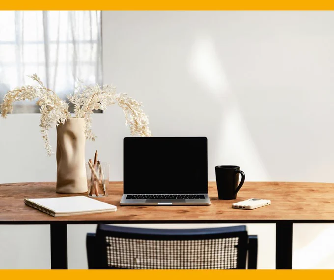 A laptop on a wooden desk in a white room. There is a notepad, vase with flowers and mug beside it