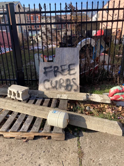 Iron fence along a concrete sidewalk on a sunny day. Black spray-painted board leaning against fence reads “free curbs.” Two concrete and rebar curbs sit on palettes, about 5 ft long.
