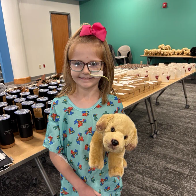 McKinley holding a stuffed dog toy, standing in front of a table with various jars and tumbler cups, wearing a blue shirt with colorful patterns and a pink bow in their hair.
