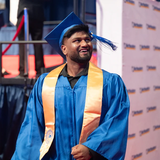 A graduate wearing blue and gold regalia smiles while looking into the stands