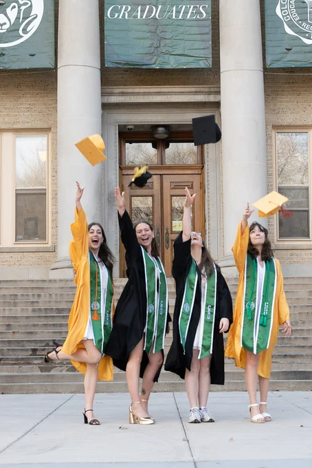 Four graduates in their graduation regalia throwing their graduation caps in the air in front of the Administration Building.