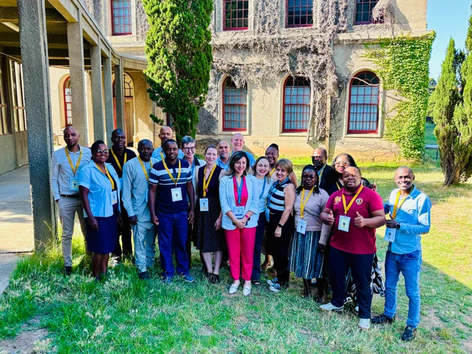 Participants of the Open Publishing workshop gather outside on a sunny day surrounded by building pillars, trees, and vines framing the windows in the background. 