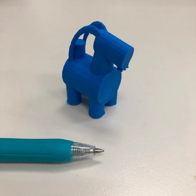 A small blue figurine in the shape of a goat similar to the large goat made of hay erected in Gavle Sweden.  A pen tip is included for a sense of scale.  The figurine is approximately 5 cm tall