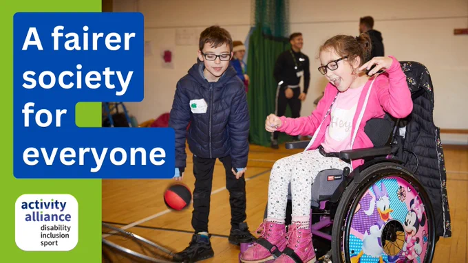 A young girl in a wheelchair and a young boy are taking part in an inclusive activity in a sports hall. They are enjoying tossing a ball through a hoop. There is also text on the picture that says: A fairer society for everyone.
