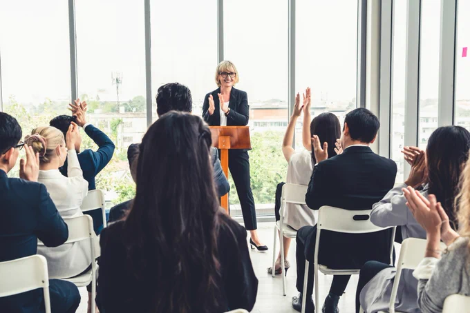 A female lecture standing in front of a room with attendees that are applauding. You see the room from behind, and the crowd is diverse with female and male attendees.