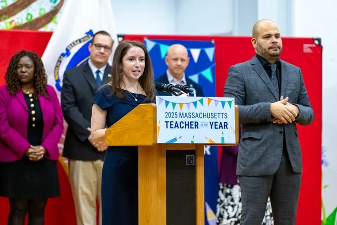Luisa Sparrow at a podium. Behind her is Boston City Council President Ruthzee Louijeune, Rep. David Biele, and Perry School Principal Andrew Rollins. A sign language interpreter is beside her.