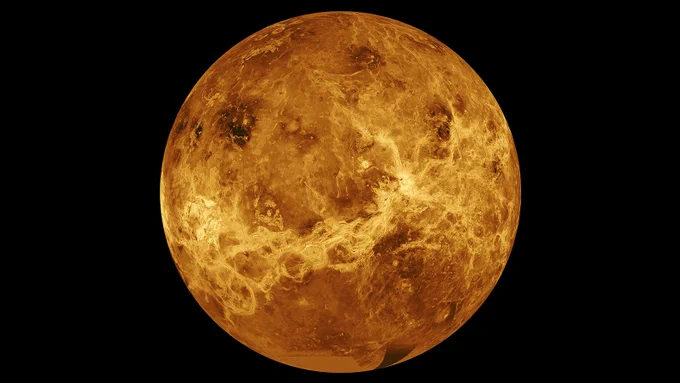 Composite image of Venus from space. The planet is a gold and dark orange mottled globe against a black background. Along the lower third, a lighter gold waving line cross from the lower left along to the center right. Darker patches appear across the upper part of the planet.