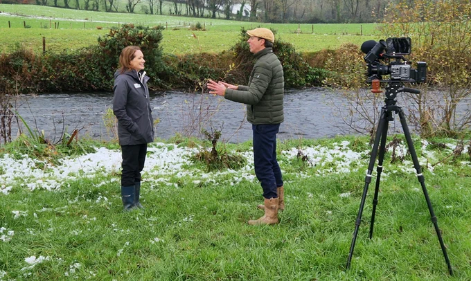 Farming For Water's Mairead Shore being interviewed by presenter Darragh McCullough beside a small river, with a video camera set up on a tripod facing them. The landscape is grassy with a hint of snow, and there are hedges in the background.