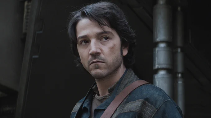A shot from the series Andor featuring Cassian Andor. Cassian is in the center looking focused, he has a bruise on his left cheek, and is wearing a blue jacket with a brown satchel .