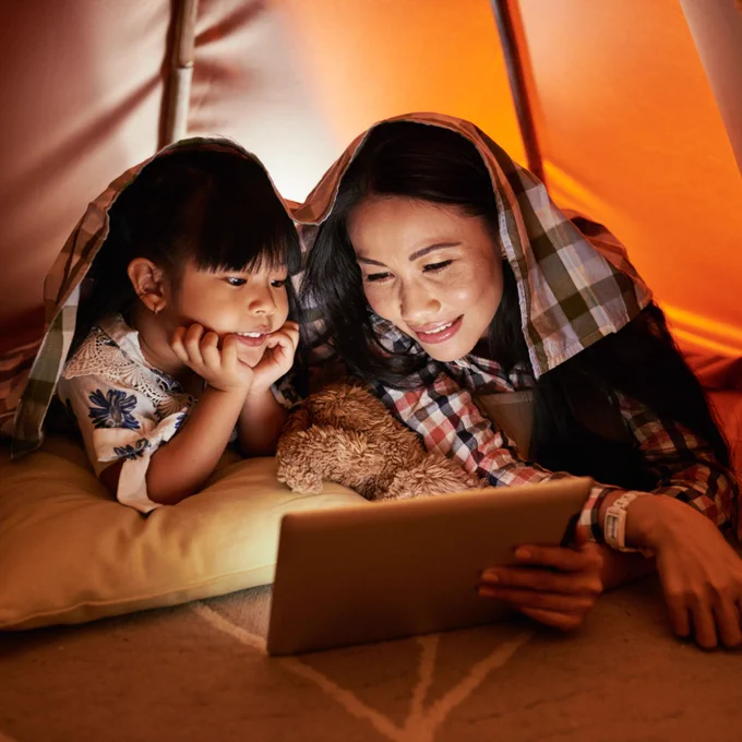 Mom and daughter lay inside a blanket tent and watch a movie on a tablet