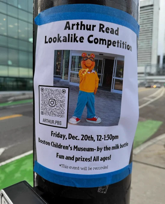 A poster is taped to a pole that reads "Arthur Read Lookalike Competition." Poster includes a picture of someone in an Arthur costume pointing at the viewer. It's happening Friday, Dec 20th from 12pm-1:30pm at the Boston Children's Museum (by the milk bottle). There will be prizes! It's for all ages. Please note the event will be recorded. 