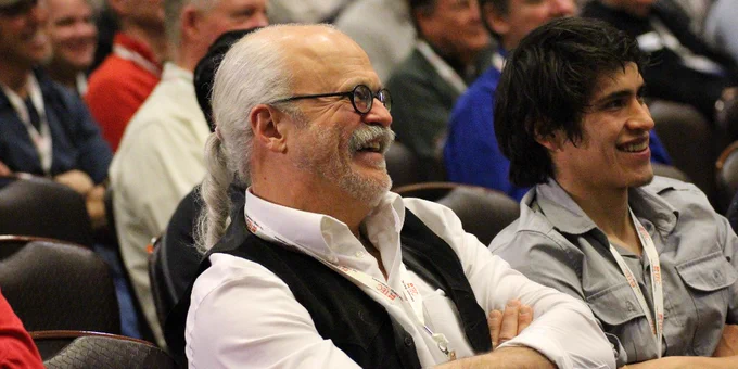 Two attendees smiling and seated at a conference, one with white hair and a vest, the other with dark hair and a light blue shirt.