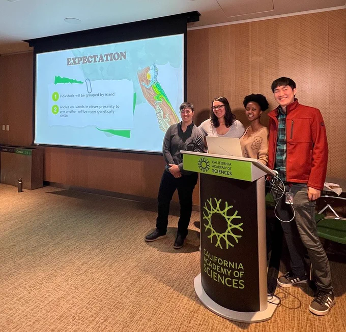 Ayanna Browne poses with her graduate committee (Dr. Lauren Esposito, Dr. Rayna Bell and Dr. Michael Yuan). A slide from her seminar presentation is in the background with an illustration of two anole species and the expected results of her analyses: 1. individuals will be grouped by island 2. anoles on islands in closer proximity to one another will be more genetically similar.