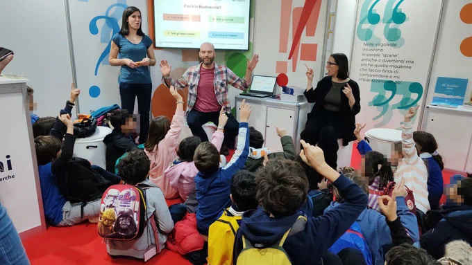 Una platea di bambini visti di spalle e seduti ascolta attentamente due persone in piedi: sono Chiara Anzolini e Fabio De Pascale, ricercatori dell'Università di Padova e del National Biodiversity Future Center oltre che curatori di The Biodiversity Issue di Comics&Science. Si trovano alla fiera della piccola e media editoria Più Libri Più Liberi di Roma.