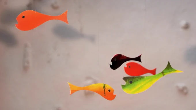 Indoors. A little fish mobile made out of colourful printed cardboard, hung on string and wire. Each fish is about 5 cm long.