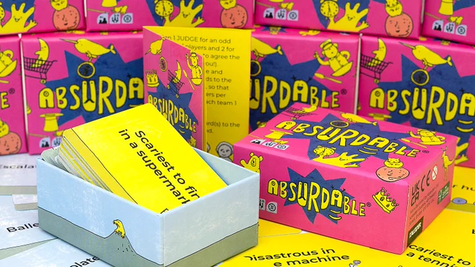 An open box of Absurdable, a card game, sitting on a table covered in cards. The box has a bright pink lid that says Absurdable in big, fun letters, and it has several funny illustrations on it. The cards are also illustrated and read things like: scariest to find in a supermarket, chocolate, disastrous in a time machine, etc.
