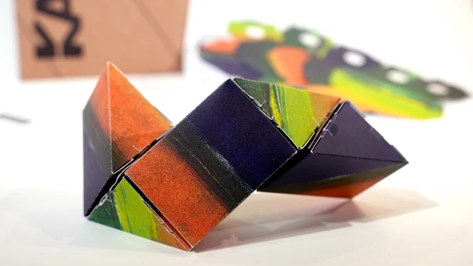A little cardboard toy made of five interconnected prisms sitting on a table. It is printed in bright bands of blue, orange and green ink.