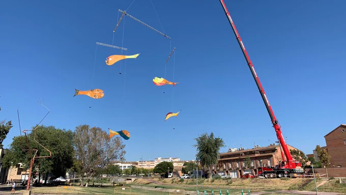 Outdoors on a sunny day, a huge crane holds a giant fish mobile about 50 metres in the air. Five fish hang from it, each one about 8 metres long. People stroll in the park underneath it, while others stop to look up at the mobile.