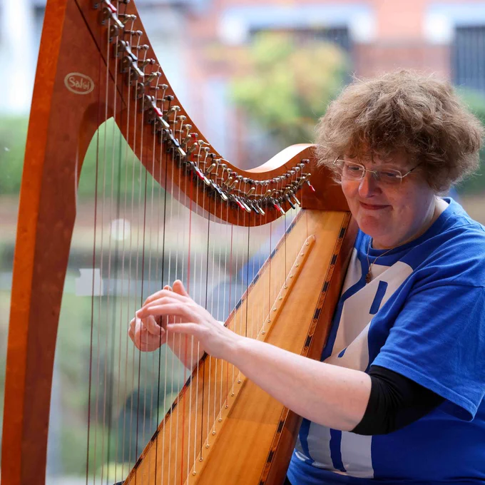 A square image showing a harpist. The harpist is wearing a blue t-shirt and holding and playing a large wooden harp. The harpist is smiling. In the background is a blurred image showing a canal and towpath landscape. 
