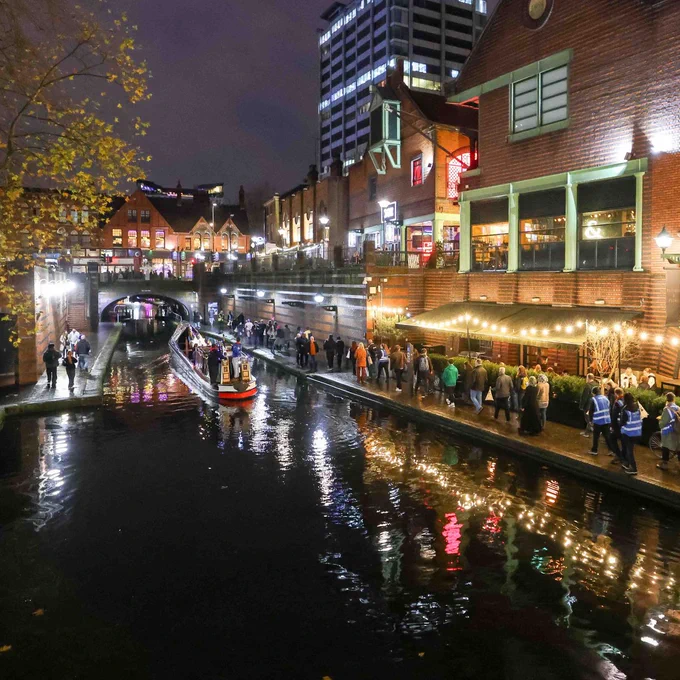 A square image showing a canal and a towpath. On the towpath is a row of shop fronts that are illuminated, and the lights are reflected in the canal below. On the towpath are a line of people carrying lanterns. On the canal is a boat. 